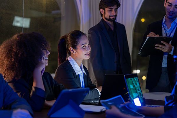 Young professionals businesswoman working together in a modern office at night, discussing ideas with laptops and digital devices in teamwork session.