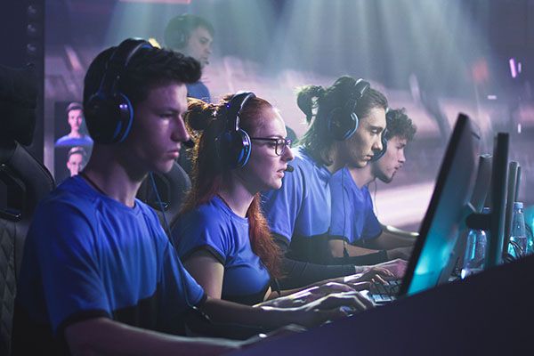 Young and successful men and women professional gamers with coach playing video game on computers against monitor with match broadcast during esports tournament