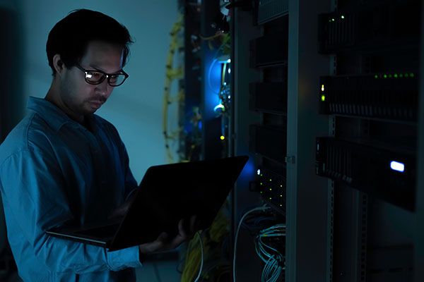 Focused Asian male IT network administrator uses a laptop to monitor server performance inside a dark data center room managing complex corporate cybersecurity operations and infrastructure.