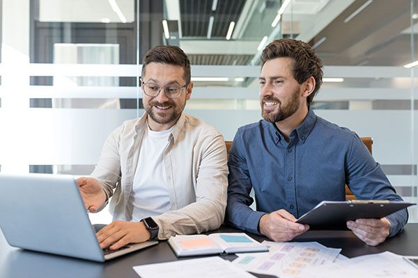 Two male business professionals are engaging in a collaborative meeting at a modern office, one pointing at a laptop screen while the other holds a clipboard, both smiling and discussing work