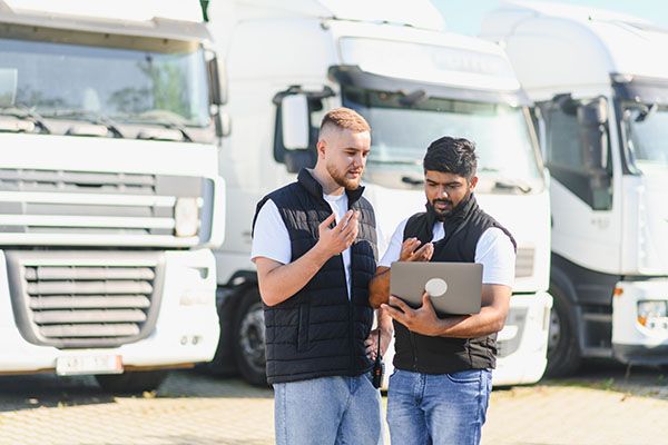 Two diverse truck drivers reviewing transport routes and cargo details on a laptop, standing by semi trucks