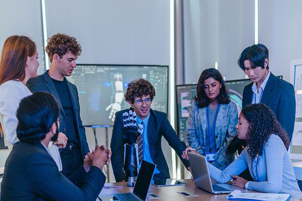 Engineers and managers in business suits gather around a conference table with a robotic arm prototype, reviewing the CAD model and test charts displayed on the meeting room screens.