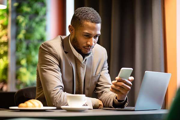 Multitasking. Confident black businessman using his mobile phone and laptop for work at coffee shop, copyspace