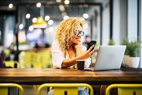 Happy young adult woman using phone and laptop at the desk in cafe with free wireless connection. Modern businesswoman working on line in coworking space. Smart people with online job indoor activity