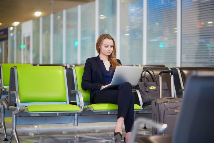 Young elegant business woman with hand luggage in international airport terminal, working on her laptop while waiting for flight