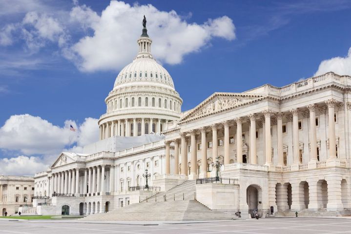 East facade of the US Capitol Building, Washington DC, USA.. American flag and vivid blue sky with clouds are in the image.