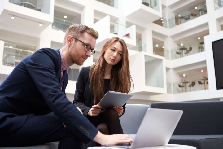 Two Businesspeople Using Laptop In Lobby Of Modern Office