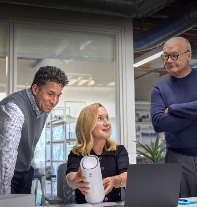 Wake Up with Craig team members (L-R: Oscar, Angela, Creed) seen from across desk. Angela seated, holding AT&T Internet Air gateway. Oscar with pleased smile, looks from behind her shoulder, Creed stands to other side with folded arms.