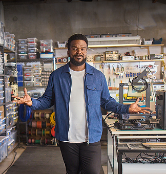 Craig team walks through his workroom with arms outstretched to show the space. The background is full of work items - contatiners, tools, wires, equipment.