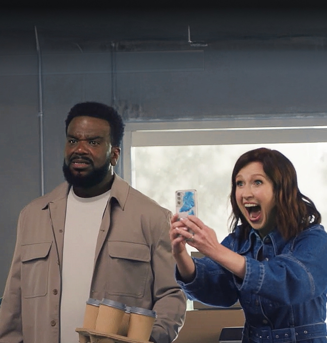 Craig standing in stunned disbelief, holding a tray of coffee cups. Ellie beside him looks delighted, phone out to record what they’re reacting to.