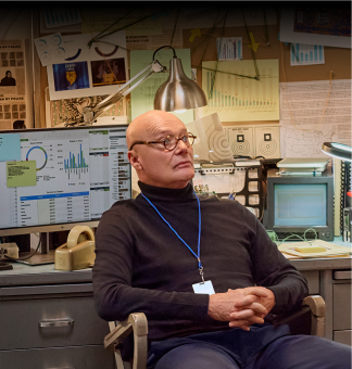 Creed is sitting in his office with chair turned around and hands folded in front of him. He is leaning back with a serious look on his face. His office has many items, including two computer monitors and a full bulletin board.