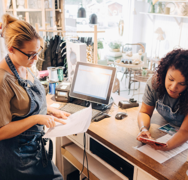 Two women working in a boutique review documents and a smartphone at the counter.
